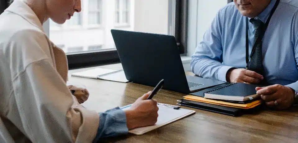 entretien-embauche-recrutement-bureau Personne écrivant sur une tablette pendant un entretien d'embauche avec un recruteur devant un ordinateur portable et des dossiers.